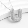 The Collar Inicial Bubble - ¡Llévate 2 por el precio de 1! es un collar plateado con colgante grande en forma de U con cristales brillantes, ideal como collar personalizado y exhibido elegantemente sobre un fondo blanco curvado.