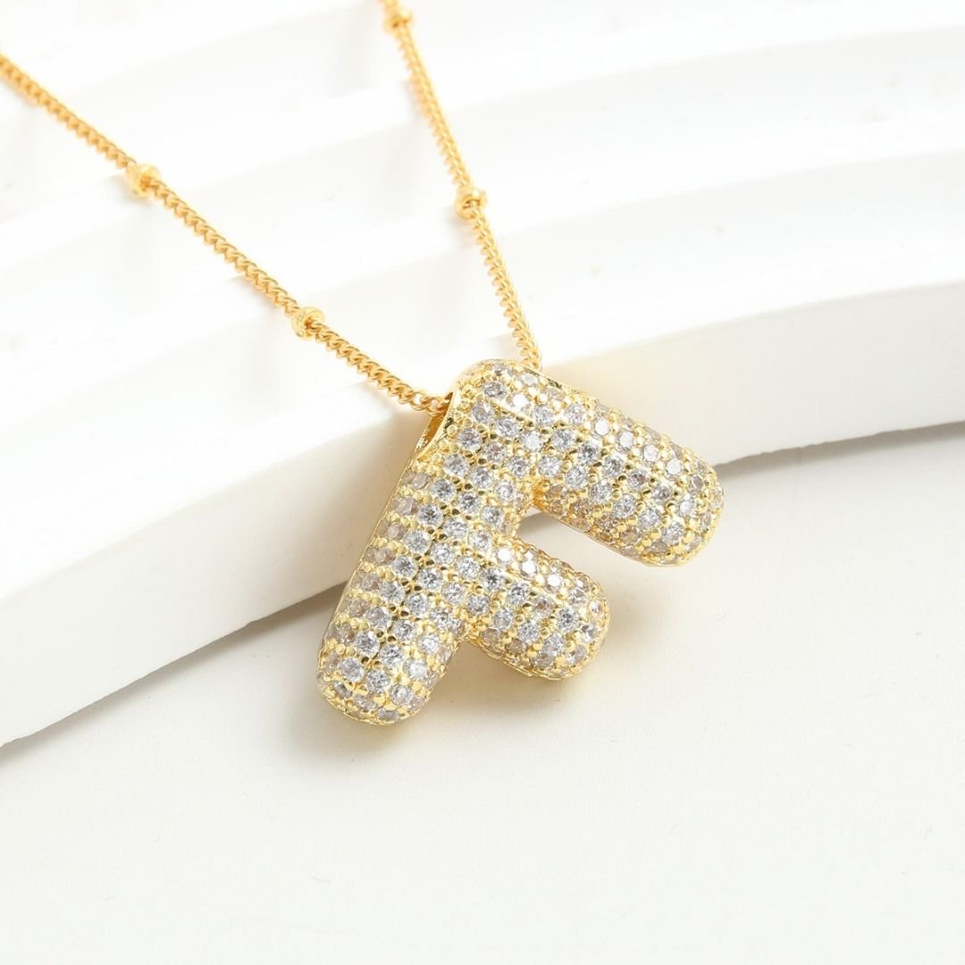 El Collar Inicial Bubble - ¡Llévate 2 por el precio de 1! en oro, con colgante de letra F y brillantes, se exhibe sobre una superficie blanca. Un regalo personalizado ideal para esa persona especial.