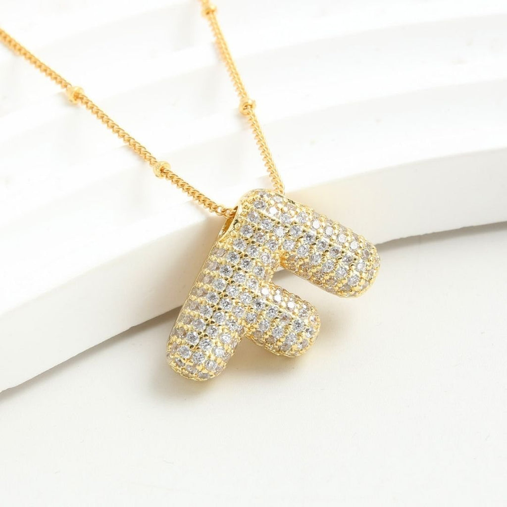 El Collar Inicial Bubble - ¡Llévate 2 por el precio de 1! en oro, con colgante de letra F y brillantes, se exhibe sobre una superficie blanca. Un regalo personalizado ideal para esa persona especial.