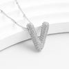 El Collar Inicial Bubble, con dije en forma de V decorado con cristales brillantes, luce elegante sobre una superficie blanca curva. ¡Aprovecha la promoción y llévate 2 por el precio de 1!.