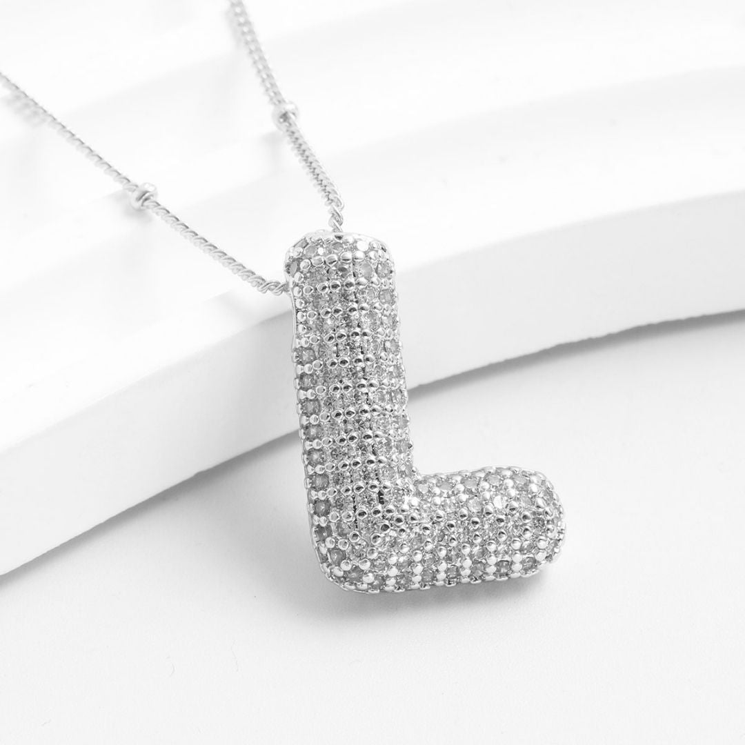 El Collar Inicial Bubble es un collar plateado con dije en forma de L decorado con brillantes, sobre fondo blanco. ¡Llévate 2 por el precio de 1 y obsequia un regalo personalizado único!.