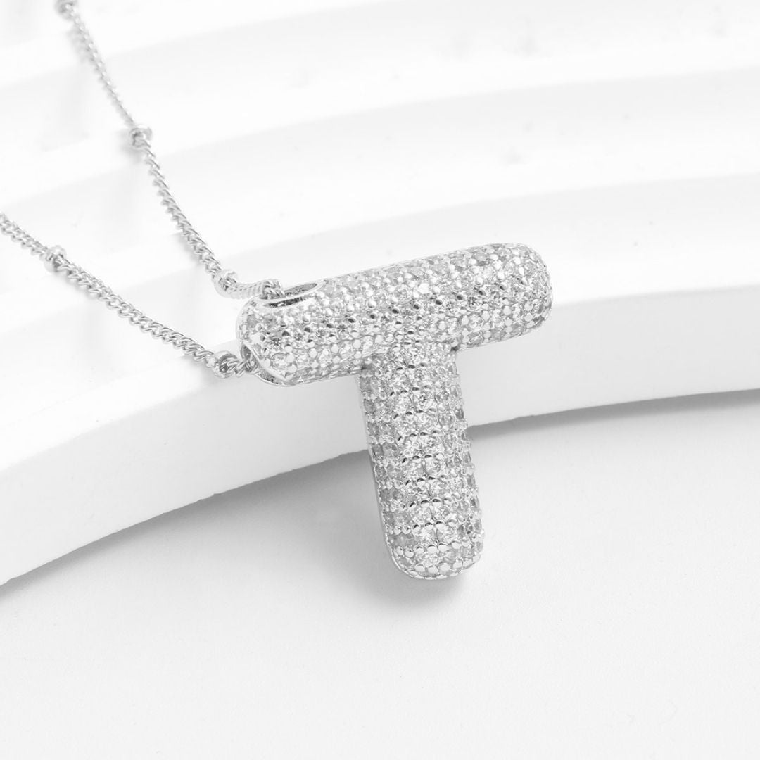 El Collar Inicial Bubble, en promoción de 2x1, es un collar plateado con dije en forma de "T" adornado de brillantes, presentado sobre un fondo blanco texturizado.