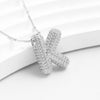 El Collar Inicial Bubble - ¡Llévate 2 por el precio de 1! es un collar plateado con dije en forma de letra K, decorado con brillantes strass, ideal como regalo personalizado y presentado sobre fondo blanco.