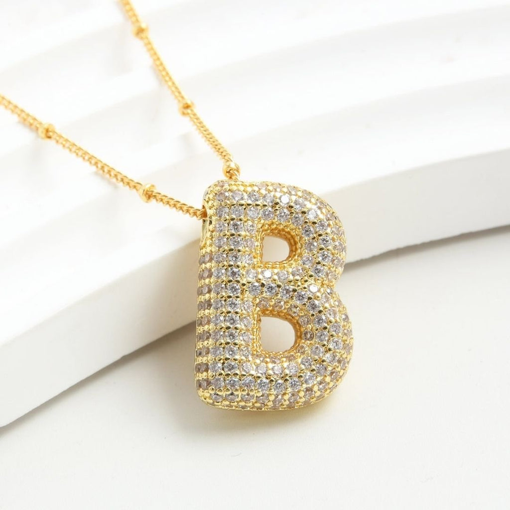 El Collar Inicial Bubble - ¡Llévate 2 por el precio de 1! es un collar dorado con dije grande de letra "B" cubierto de brillantes, exhibido sobre fondo blanco texturizado-un regalo personalizado perfecto.