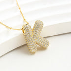 El Collar Inicial Bubble - ¡Llévate 2 por el precio de 1! es un collar dorado con inicial y brillantes, exhibido sobre fondo blanco texturizado; ideal como regalo personalizado para alguien especial.