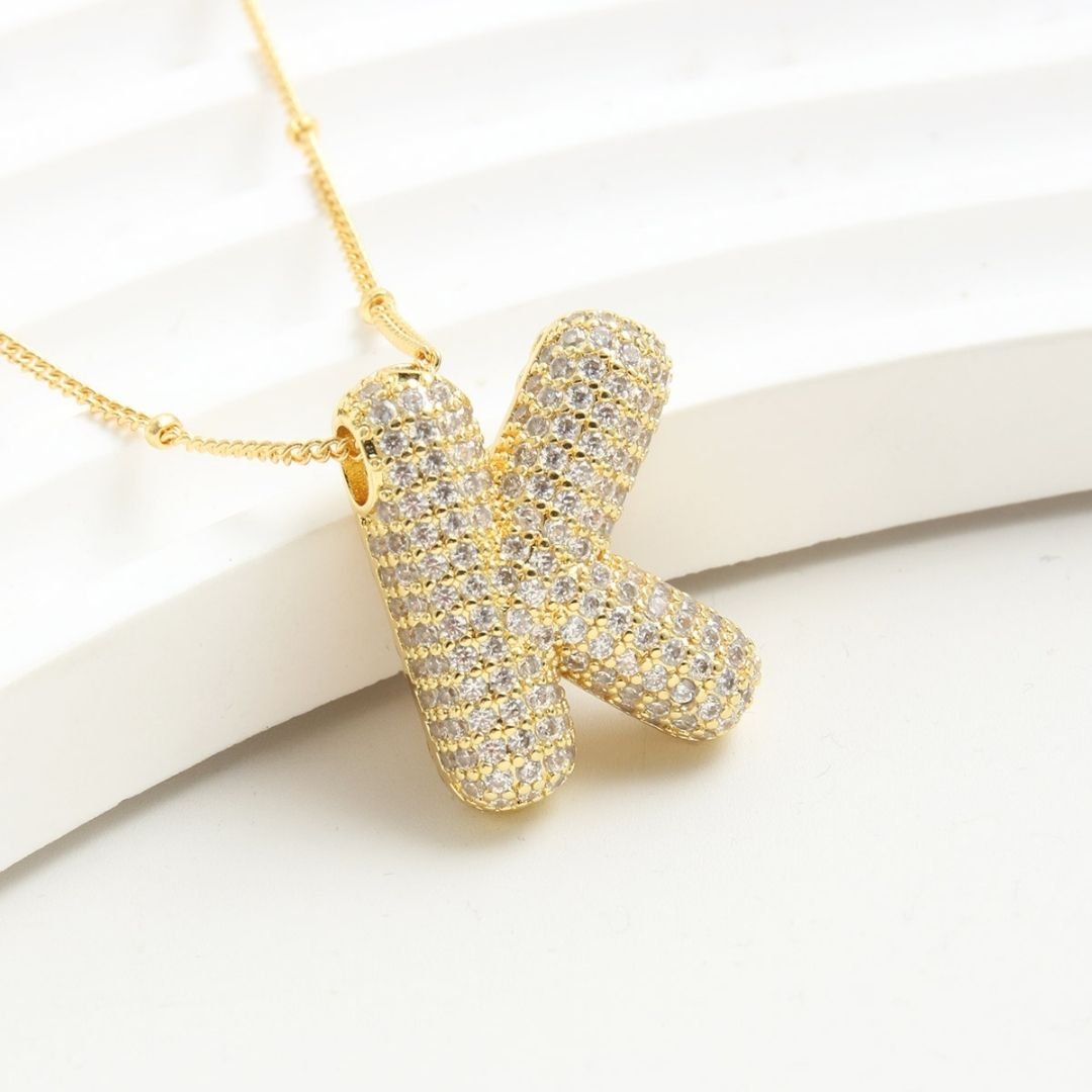 El Collar Inicial Bubble - ¡Llévate 2 por el precio de 1! es un collar dorado con inicial y brillantes, exhibido sobre fondo blanco texturizado; ideal como regalo personalizado para alguien especial.