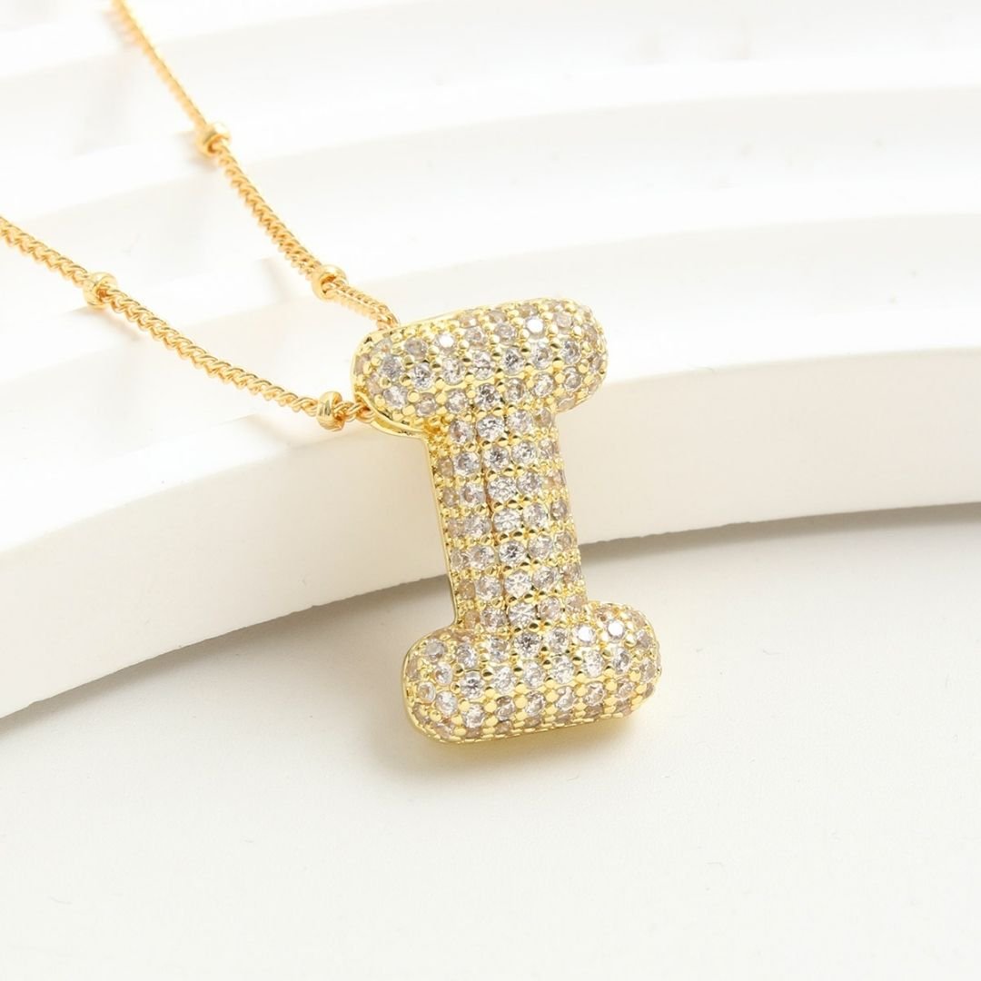The Collar Inicial Bubble - ¡Llévate 2 por el precio de 1! es un collar dorado con dije de inicial, incrustado con cristales claros sobre fondo blanco texturizado, ideal como collar personalizado o regalo especial.