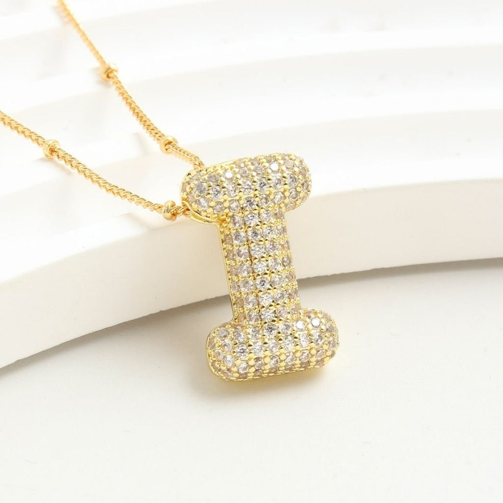 The Collar Inicial Bubble - ¡Llévate 2 por el precio de 1! es un collar dorado con dije de inicial, incrustado con cristales claros sobre fondo blanco texturizado, ideal como collar personalizado o regalo especial.