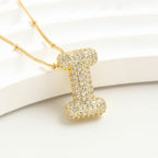 The Collar Inicial Bubble - ¡Llévate 2 por el precio de 1! es un collar dorado con dije de inicial, incrustado con cristales claros sobre fondo blanco texturizado, ideal como collar personalizado o regalo especial.