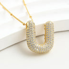 El Collar Inicial Bubble - ¡Llévate 2 por el precio de 1! es un collar de cadena de oro con un gran colgante en forma de U incrustado de brillantes piedras blancas, que se muestra sobre un fondo blanco.