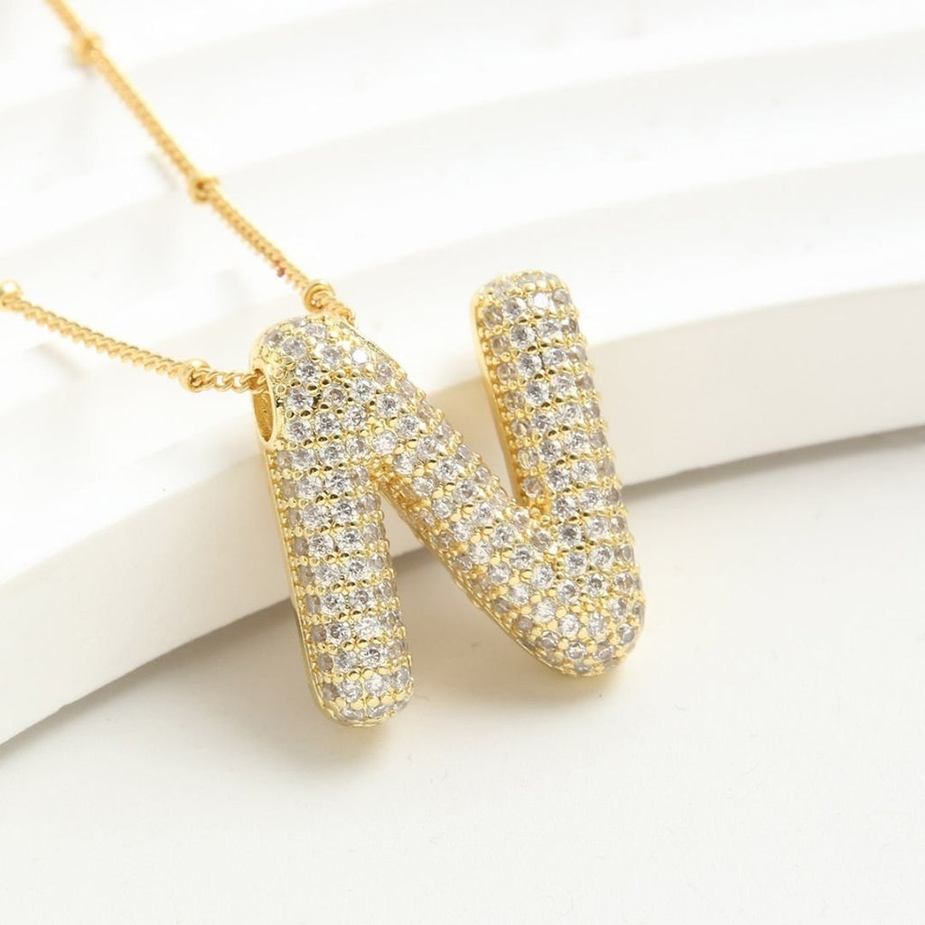 Collar Inicial Bubble: cadena dorada con colgante en forma de letra N, decorado con piedras blancas brillantes. ¡Llévate 2 por el precio de 1! Ideal como regalo personalizado. Exhibido sobre una superficie blanca.