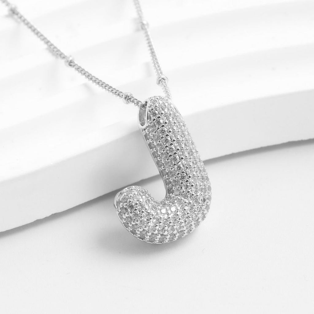 El Collar Inicial Bubble, con dije en forma de letra J y cristales brillantes, se muestra sobre una superficie blanca texturizada. ¡Llévate 2 por el precio de 1 y obtén el regalo personalizado perfecto!.