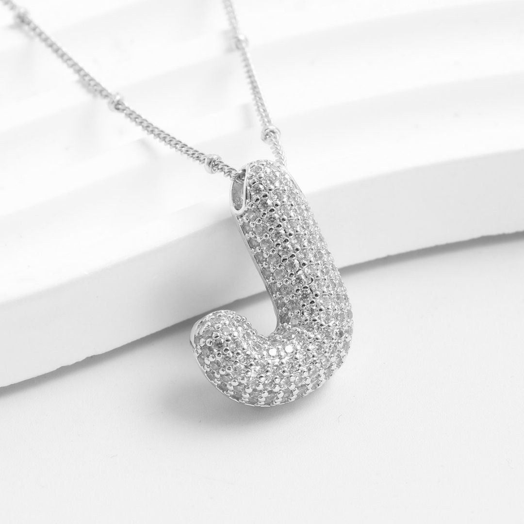 El Collar Inicial Bubble, con dije en forma de letra J y cristales brillantes, se muestra sobre una superficie blanca texturizada. ¡Llévate 2 por el precio de 1 y obtén el regalo personalizado perfecto!.