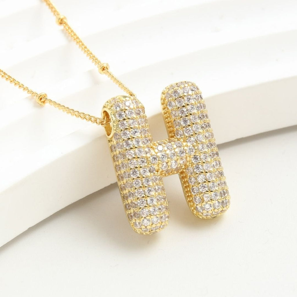 El Collar Inicial Bubble (¡Llévate 2 por el precio de 1!) es un collar dorado con dije en forma de H, decorado con brillantes piedras claras, ideal como regalo personalizado y presentado sobre fondo blanco.