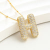 El Collar Inicial Bubble (¡Llévate 2 por el precio de 1!) es un collar dorado con dije en forma de H, decorado con brillantes piedras claras, ideal como regalo personalizado y presentado sobre fondo blanco.