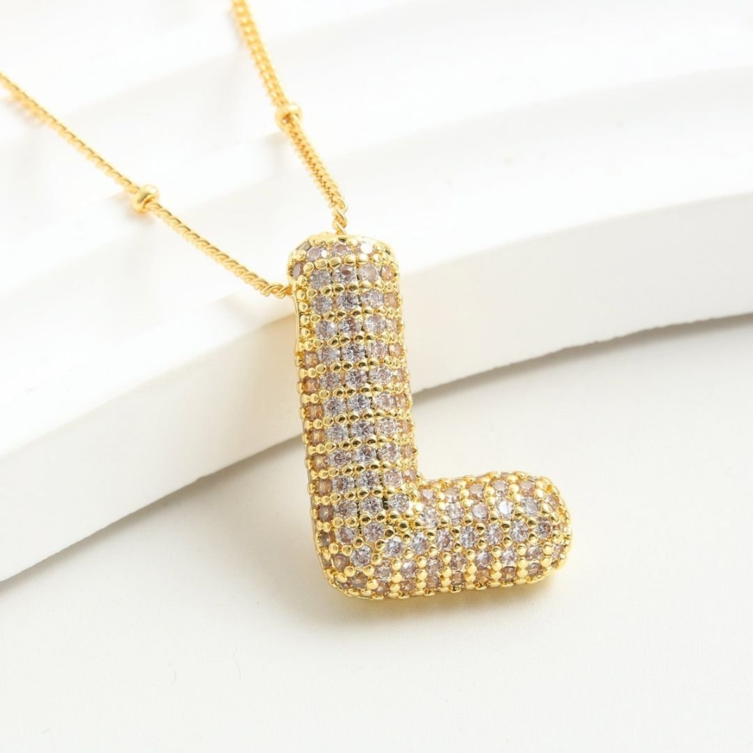Collar Inicial Bubble - ¡Lévate 2 por el precio de 1! Un collar dorado con dije en forma de letra L y relucientes piedras, elegante sobre fondo blanco. Ideal como regalo personalizado.