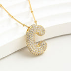 The Collar Inicial Bubble - ¡Llévate 2 por el precio de 1! es un collar dorado con dije de letra C en relieve y cristales brillantes, exhibido sobre una superficie curva blanca.