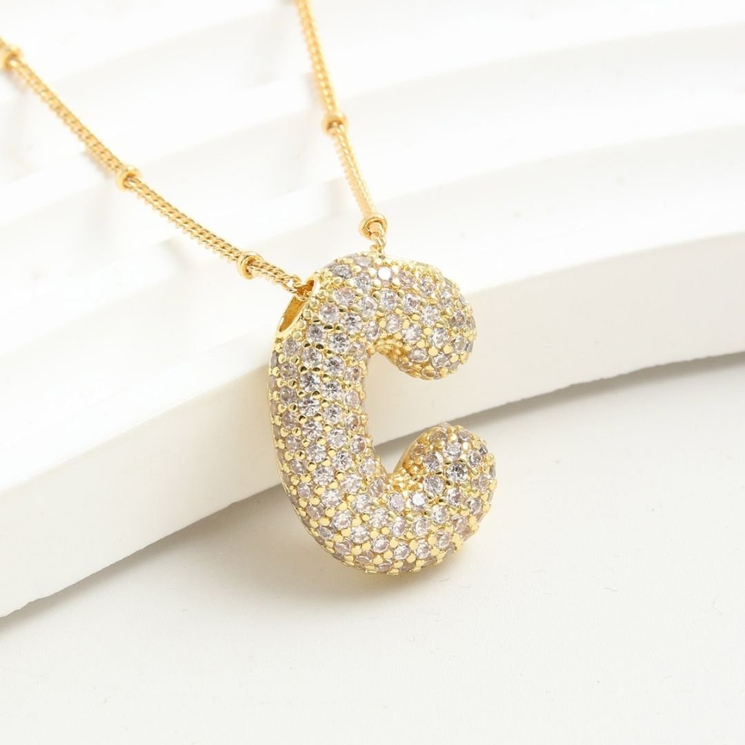 The Collar Inicial Bubble - ¡Llévate 2 por el precio de 1! es un collar dorado con dije de letra C en relieve y cristales brillantes, exhibido sobre una superficie curva blanca.