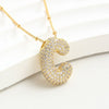 The Collar Inicial Bubble - ¡Llévate 2 por el precio de 1! es un collar dorado con dije de letra C en relieve y cristales brillantes, exhibido sobre una superficie curva blanca.
