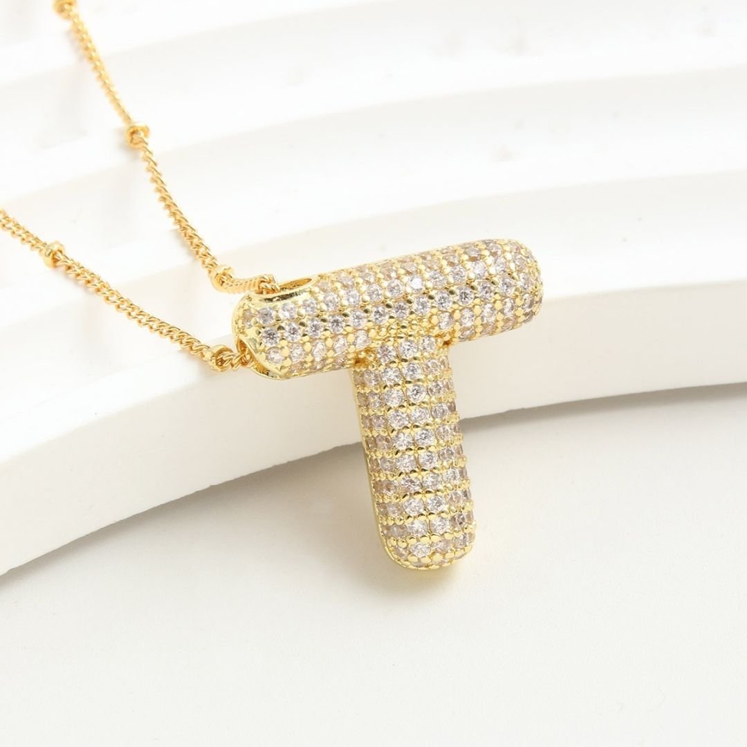 El Collar Inicial Bubble - ¡Llévate 2 por el precio de 1! destaca con su inicial dorada y diamantes brillantes, siendo un regalo personalizado ideal que resalta sobre un fondo blanco texturizado.