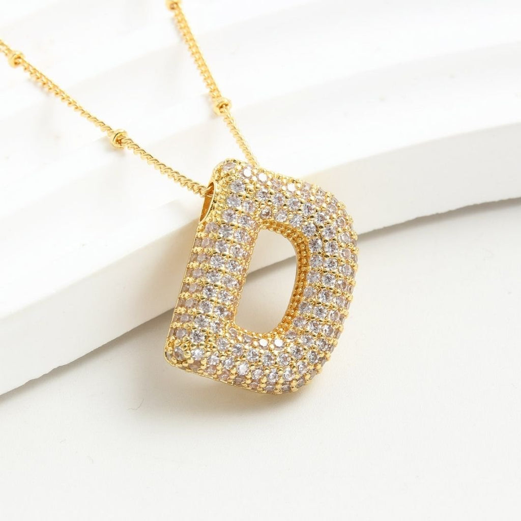 A gold "Collar Inicial Bubble" con un gran dije letra D adornado con piedras brillantes, sobre fondo blanco. ¡Llévate 2 por el precio de 1! Ideal como regalo personalizado.