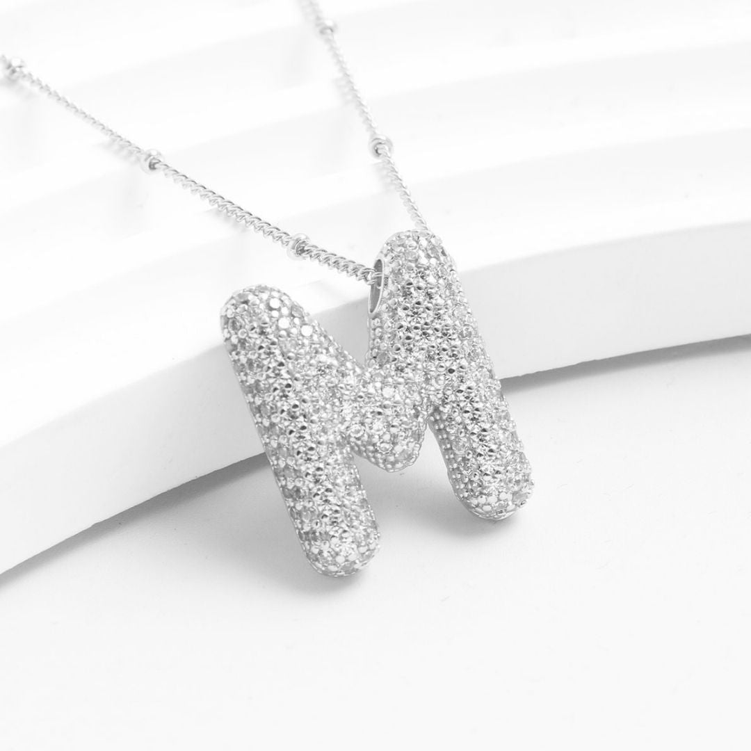 El collar Inicial Bubble, que se muestra en plata con un brillante colgante de una M de strass sobre fondo blanco, es ideal como collar personalizado o regalo personalizado-¡llévate 2 por el precio de 1!.