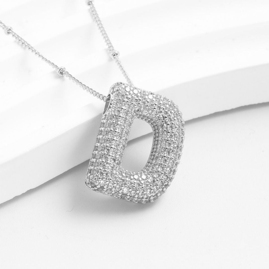 El Collar Inicial Bubble, bañado en plata y cubierto de brillantes cristales, se exhibe sobre una superficie blanca curva; ideal como regalo personalizado. ¡Llévate 2 por el precio de 1!.