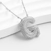 The Collar Inicial Bubble - ¡Llévate 2 por el precio de 1! es un collar plateado con una gran letra G texturizada y adornada con diamantes, presentado elegantemente sobre una superficie blanca.