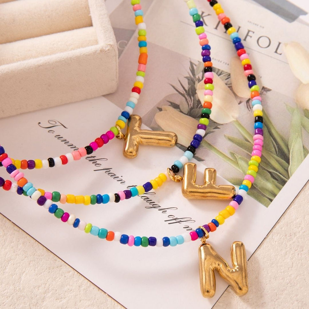 Collar Inicial colorido - ¡Lévate 3 por el precio de 1! Coloridas cuentas con dijes dorados de letras "Y", "L" y "N", exhibidos sobre una superficie crema cerca de una bandeja y revista floral-un regalo ideal para amigas o para ti.