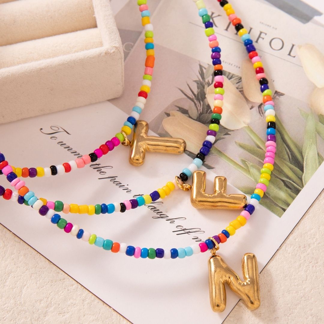 Collar Inicial colorido - ¡Lévate 3 por el precio de 1! Coloridas cuentas con dijes dorados de letras "Y", "L" y "N", exhibidos sobre una superficie crema cerca de una bandeja y revista floral-un regalo ideal para amigas o para ti.