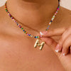 Una persona luce el Collar Inicial colorido - ¡Llévate 3 por el precio de 1!, un collar con cuentas coloridas y un gran dije dorado con la letra M, sosteniéndolo entre sus dedos mientras muestra una manicura francesa.