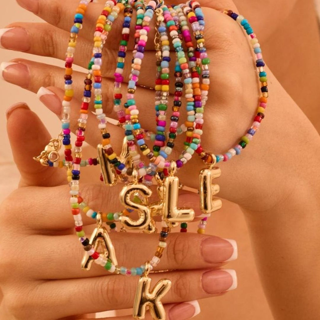 Una mano con manicura francesa sostiene varios "Collar Inicial colorido - ¡Llévate 3 por el precio de 1!" con cuentas de colores y dijes dorados en forma de letras (M, S, L, E, K) - ideales como regalo para amigas o un collar personalizado.