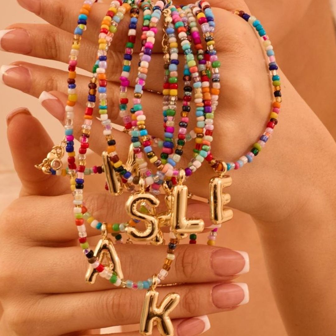 Una mano con manicura francesa sostiene varios "Collar Inicial colorido - ¡Llévate 3 por el precio de 1!" con cuentas de colores y dijes dorados en forma de letras (M, S, L, E, K) - ideales como regalo para amigas o un collar personalizado.