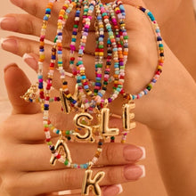 Cargar imagen en el visor de la galería, Una mano con manicura francesa sostiene varios &quot;Collar Inicial colorido - ¡Llévate 3 por el precio de 1!&quot; con cuentas de colores y dijes dorados en forma de letras (M, S, L, E, K) - ideales como regalo para amigas o un collar personalizado.