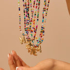 Una mano se acerca a varios collares inicial coloridos, cada uno con un dije dorado de letra, sobre fondo neutro. El Collar Inicial colorido - ¡Llévate 3 por el precio de 1! es ideal como regalo para amigas o collar personalizado.