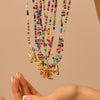 Una mano se acerca a varios collares inicial coloridos, cada uno con un dije dorado de letra, sobre fondo neutro. El Collar Inicial colorido - ¡Llévate 3 por el precio de 1! es ideal como regalo para amigas o collar personalizado.