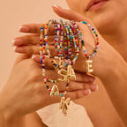 Una mujer con uñas cuidadas sostiene varios Collar Inicial colorido, cada uno con gran dije dorado de letra-¡llévate 3 por el precio de 1!-ideal como collar personalizado o regalo para amigas, sobre fondo neutro.
