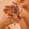 Una mujer con uñas cuidadas sostiene varios Collar Inicial colorido, cada uno con gran dije dorado de letra-¡llévate 3 por el precio de 1!-ideal como collar personalizado o regalo para amigas, sobre fondo neutro.