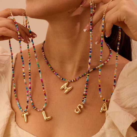 Una mujer luce varios "Collar Inicial colorido - ¡Llévate 3 por el precio de 1!", cada uno con una inicial dorada (T, L, M, S y A). Son el regalo ideal para amigas. Ella lleva un outfit claro sobre fondo neutro.