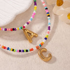 Dos collares inicial coloridos con dijes dorados de la letra A y C reposan junto a una concha y flores blancas-el Collar Inicial colorido, ¡llévate 3 por el precio de 1! Perfectos como un collar personalizado o regalo para amigas.