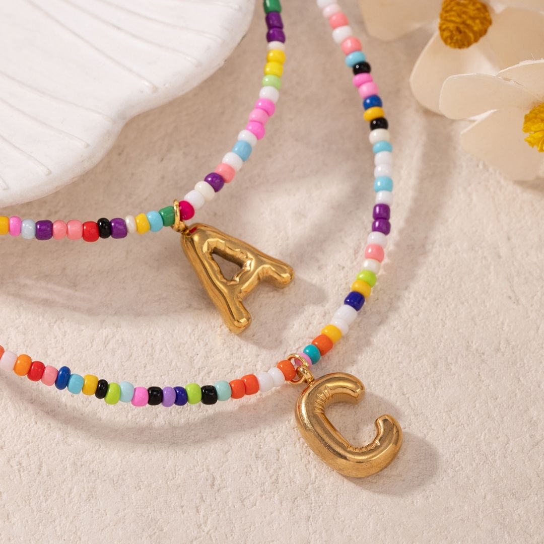 Dos collares inicial coloridos con dijes dorados de la letra A y C reposan junto a una concha y flores blancas-el Collar Inicial colorido, ¡llévate 3 por el precio de 1! Perfectos como un collar personalizado o regalo para amigas.