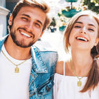 Un hombre sonriente con una chaqueta vaquera y una mujer con un top blanco sin hombros están uno al lado del otro al aire libre, ambos con el collar IZAC con inicial personalizado con colgantes rectangulares, disfrutando de un día soleado.