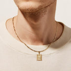 Primer plano de una persona con camisa color crema y dos collares de oro, entre ellos el Collar IZAC con inicial personalizada con colgante rectangular y diseño intrincado. Sólo son visibles la parte inferior de la cara y el cuello.