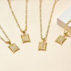 Cinco collares IZAC de oro con colgantes cuadrados ornamentados con las iniciales M, J, K y N grabadas se disponen sobre una superficie de color beige claro junto a una caja blanca-ideal como Collar IZAC con inicial personalizada.