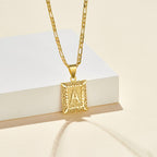 El collar IZAC con inicial personalizada es un collar de oro con un colgante rectangular de filigrana con la letra A, que aparece sobre un fondo crema junto a un bloque rectangular blanco.