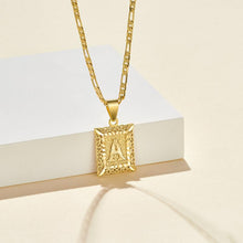 Cargar imagen en el visor de la galería, El collar IZAC con inicial personalizada es un collar de oro con un colgante rectangular de filigrana con la letra A, que aparece sobre un fondo crema junto a un bloque rectangular blanco.