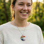 Una mujer sonríe al aire libre bajo la suave luz del sol, con un jersey claro y el collar "Luna mística" con piedras naturales y un colgante Árbol de la Vida. Los árboles verdes se difuminan suavemente en el fondo.