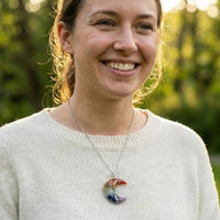 Una mujer sonríe al aire libre bajo la suave luz del sol, con un jersey claro y el collar "Luna mística" con piedras naturales y un colgante Árbol de la Vida. Los árboles verdes se difuminan suavemente en el fondo.