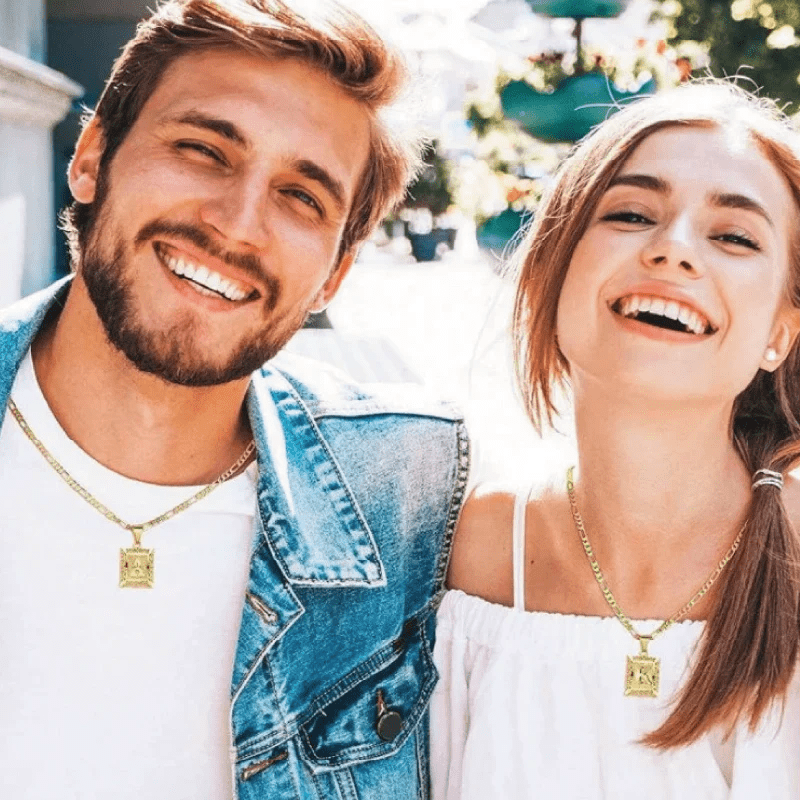 Un hombre sonriente con una chaqueta vaquera y una mujer con un top blanco sin hombros están juntos al aire libre, ambos llevan Collar Marco con inicial personalizada (Oferta 2x1), collares rectangulares de oro con colgante de acero inoxidable.