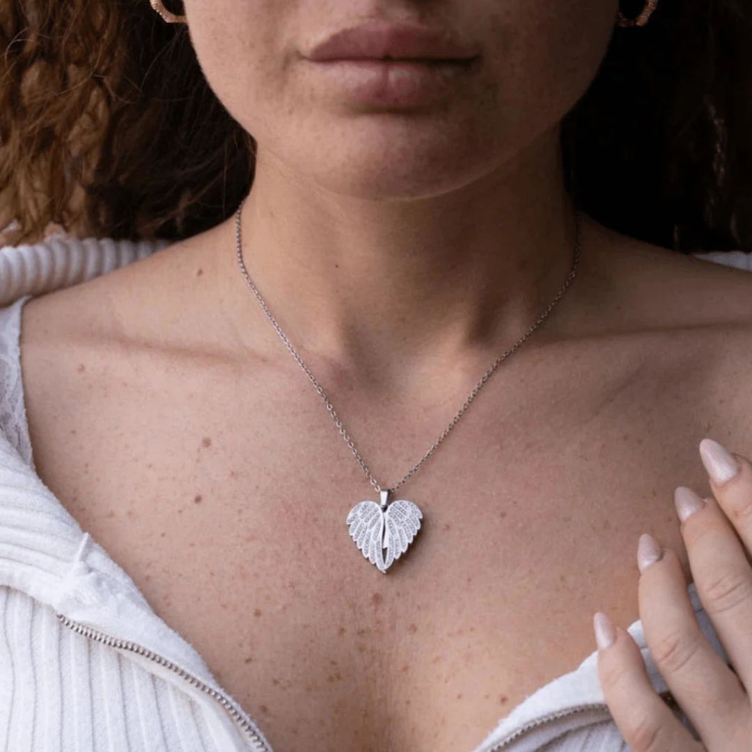 Una mujer con una camiseta blanca con cremallera lleva el collar personalizable "Mi ángel guardián" (Oferta 2X1), que incluye un colgante en forma de corazón con un diseño en forma de hoja. Su mano descansa suavemente cerca del colgante sobre su pecho.